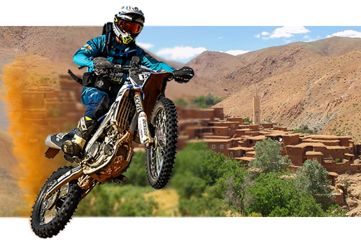 Motard en tout-terrain sur une piste de l'Atlas marocain, devant un village en terre